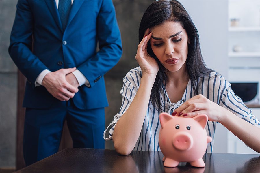 Collection Agency Bonds - View of a Worried Woman Sitting at a Table with a Piggy Bank as a Collection Agent in a Suit Stands Behind Her